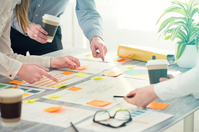 A group of people around a table with papers, charts, and sticky notes, holding coffee cups and pens, with a plant in the background.