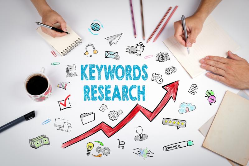 A desk with two people writing, surrounded by a notebook, coffee cup, pens, and paperclips. The center features "KEYWORDS RESEARCH" in blue text with a red upward arrow, accompanied by icons like money, a globe, gears, and a checklist, symbolizing growth and strategy.